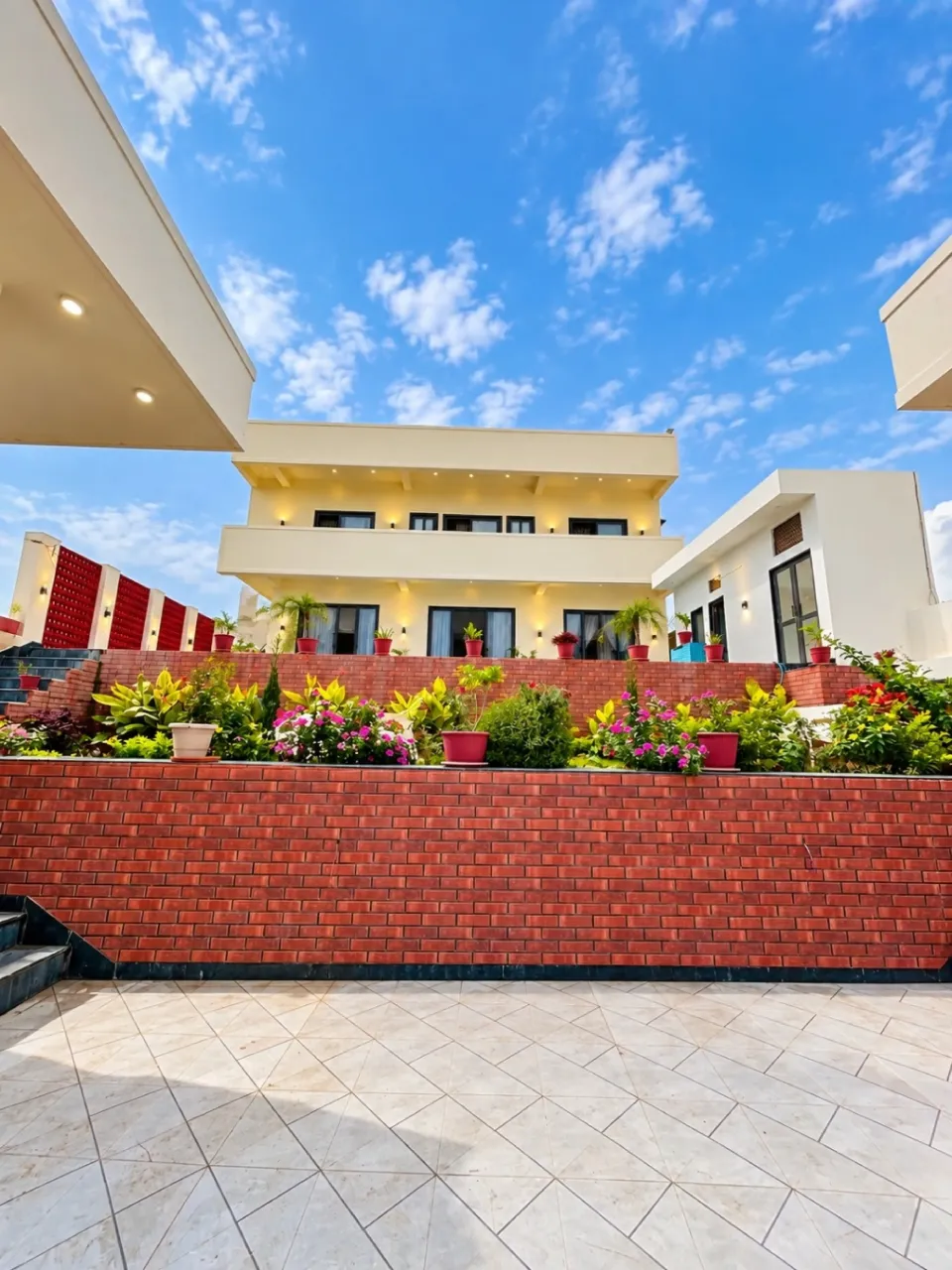 Modern 5 bhk villa with a landscaped garden, red brick walls, and a spacious tiled courtyard under blue skies